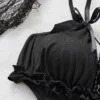 Black cut-out teddy bodysuit with ruffle-trim edges, bow-tied shoulder straps and decorative bead chains, laid flat on white backdrop with roses and Halloween accents.