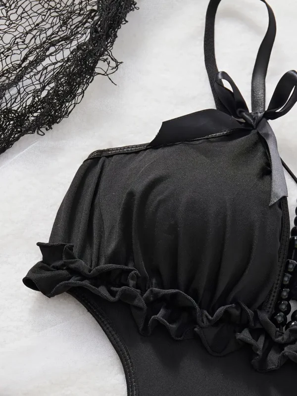 Black cut-out teddy bodysuit with ruffle-trim edges, bow-tied shoulder straps and decorative bead chains, laid flat on white backdrop with roses and Halloween accents.
