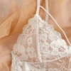 White floral lace and mesh teddy bodysuit with scalloped edges and adjustable spaghetti straps, styled on beige fabric with a red rose and vintage book page.