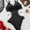 Black cut-out teddy bodysuit with ruffle-trim edges, bow-tied shoulder straps and decorative bead chains, laid flat on white backdrop with roses and Halloween accents.