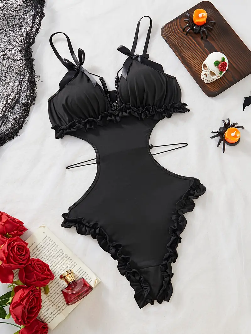 Black cut-out teddy bodysuit with ruffle-trim edges, bow-tied shoulder straps and decorative bead chains, laid flat on white backdrop with roses and Halloween accents.