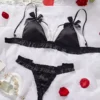 aa9af471-39b4-4e77-bfc0-73ed3922274b Black satin ruffle-trim bralette and ruched thong lingerie set with delicate pearl-chain connector and bow-topped adjustable straps, styled on white backdrop with rose petals and pearls.